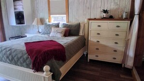 Une chambre à coucher comprenant un lit, une commode, un ventilateur de plafond, une fenêtre et une étagère fixée au mur.