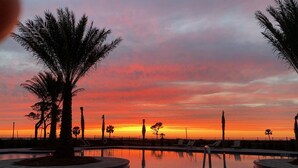 A swimming pool with palm trees and lounge chairs, surrounded by a fence, during sunset.