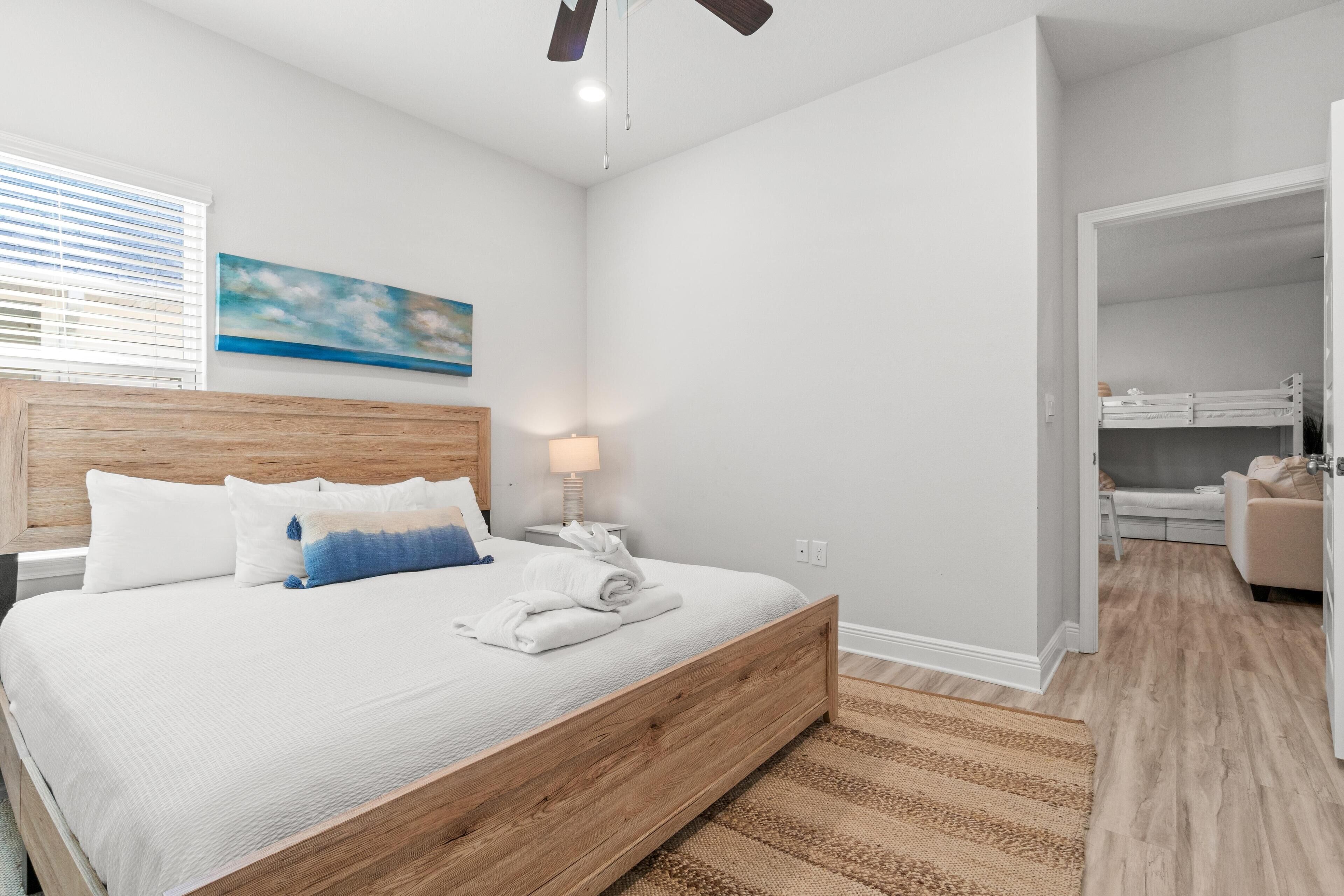 A bedroom with a wooden bed, white bedding, a nightstand, a lamp, a painting on the wall, and a view into an adjacent room.