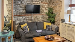 Ein Wohnzimmer mit Steinwand, einem Flachbildfernseher, einem grauen Sofa mit gelben Kissen, einem kleinen Tisch mit Fernbedienung und einem Fenster mit Jalousien.