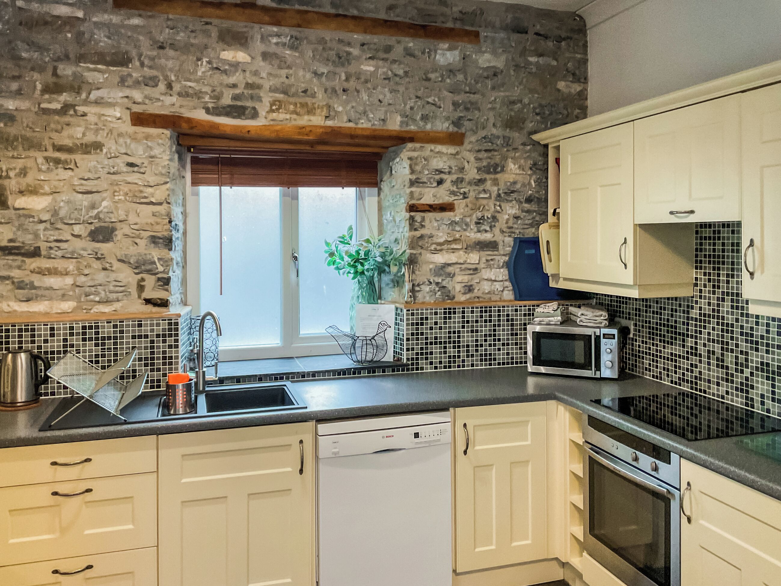 A kitchen with stone walls, a window, a sink, a microwave, and cabinets.