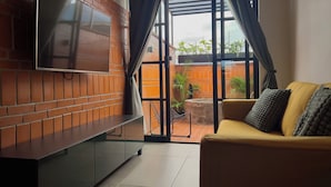 Ein Wohnzimmer mit an einer Ziegelwand montiertem Fernseher, einem gelben Sofa mit Kissen und Blick auf eine Ziegelwand sowie Pflanzen draußen.