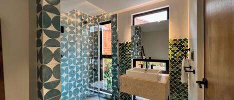 A modern bathroom with a geometric tile pattern, a glass shower enclosure, a sink with a mirror above, and a view of the outdoors.