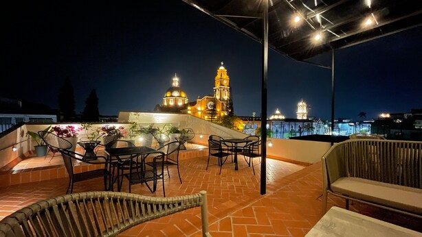 Eine Terrasse im Freien mit Korbmöbeln und Topfpflanzen, mit Blick auf eine Skyline mit beleuchteten Gebäuden und einem Uhrturm.