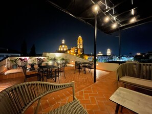An outdoor patio with wicker furniture and potted plants, overlooking a cityscape with illuminated buildings and a clock tower. - Estacion Alameda Downtown (Orizaba)