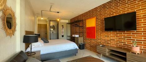 A modern hotel room with a brick wall, a flat-screen TV, a bed with white linens, a sofa, a coffee table, and a TV stand with a plant.