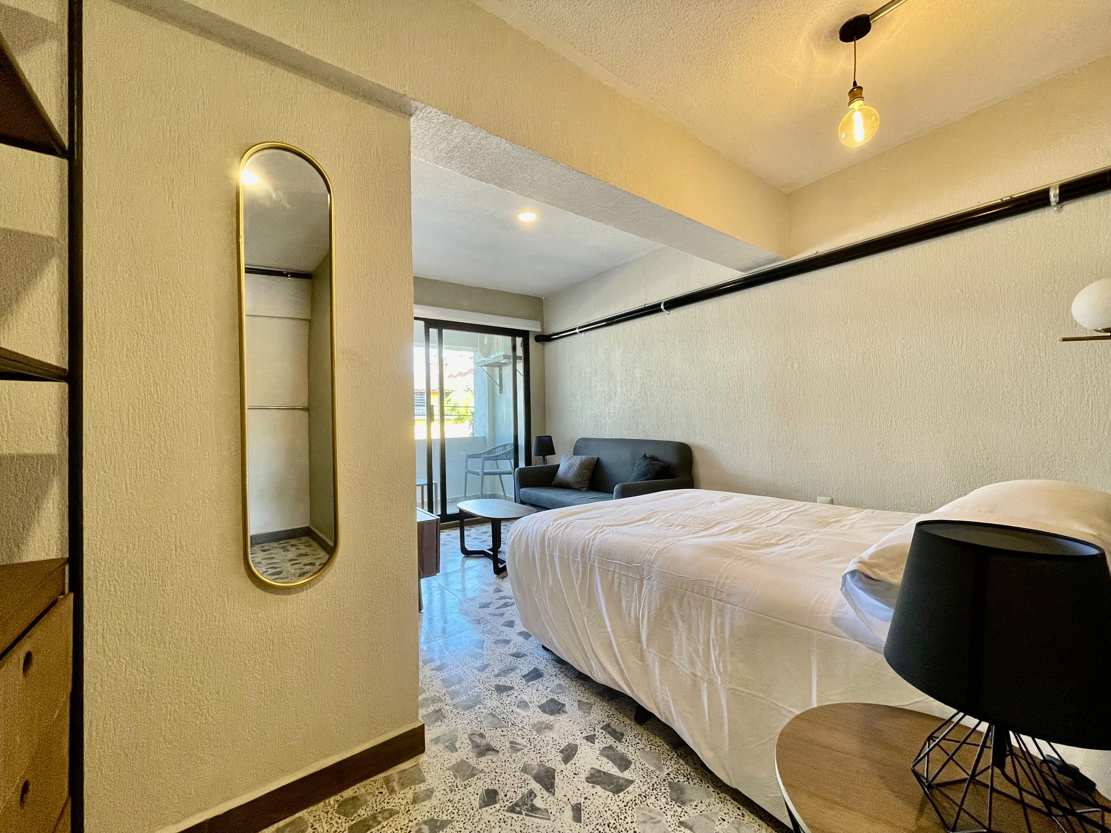 A hotel room with a bed, a desk, a chair, a mirror, and a view of the outside balcony.