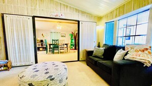 A living room with a dark-colored sofa, a round ottoman with a beach-themed cover, a dining area with a table and chairs, and a view through a door to a kitchen area. - Charming 2/2 waterfront farm home with tons of TV stations, WiFi, dock, & more (Spring Hill)