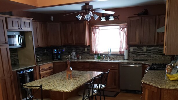 A kitchen with wooden cabinets, granite countertops, a central island with stools, a ceiling fan, and a window with curtains. - Charming Country farmhouse (Fredonia)