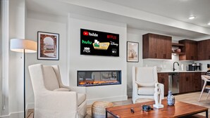 A modern living room with a fireplace, a flat-screen TV displaying streaming services, and a kitchen area with wooden cabinets.