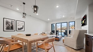 A modern living room with a wooden dining table, beige sofa, and a TV mounted on the wall.
