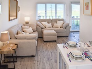 A living room with two sofas, a dining table set with plates and cutlery, a lamp, and a large window with a view of a balcony. - Lovely modern chalet with stunning sea views on a lovely quiet site. (Bacton)