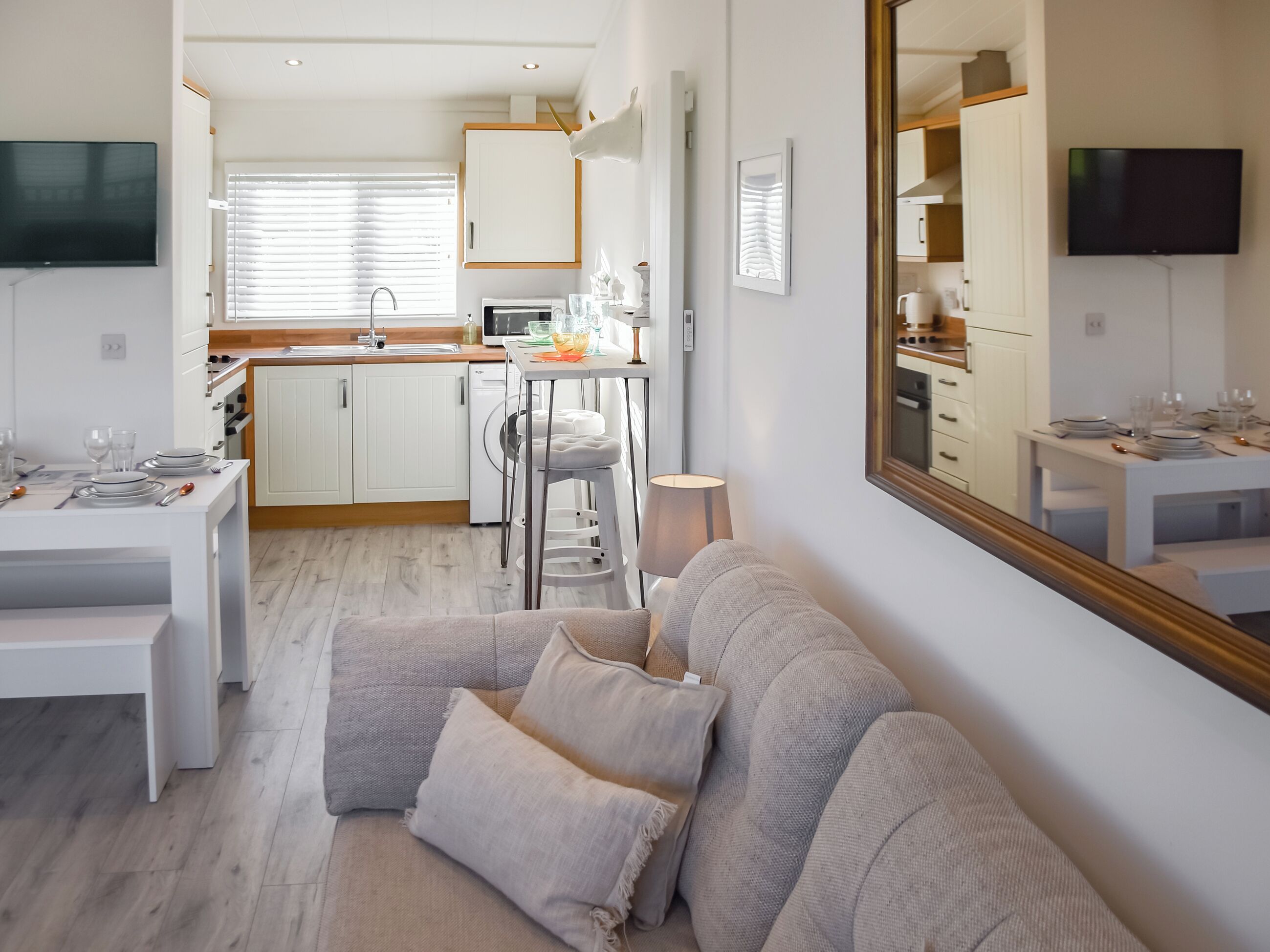 Uma sala de estar moderna com sofá, uma mesa de jantar posta para dois e uma cozinha com armários brancos e micro-ondas.
