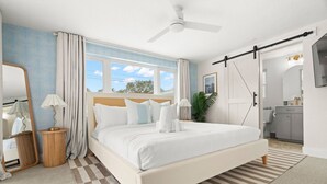 Een slaapkamer met een groot bed, een plafondventilator, een raam met gordijnen, een nachtkastje met een lamp, en een badkamer zichtbaar door een geopende deur.