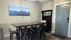 A dining area with a long wooden table and black chairs, a painting of a bridge, a black bench, and a gray door.
