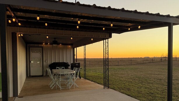 An outdoor patio with a table and chairs, a barbecue grill, and a view of a grassy field at sunset.