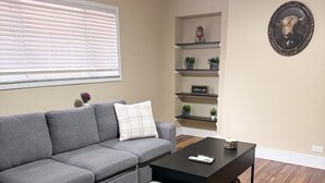 Un salon avec un canapé d’angle gris, une table basse et un ventilateur de plafond. On y trouve des étagères ornées d’objets décoratifs et un tableau encadré accroché au mur.