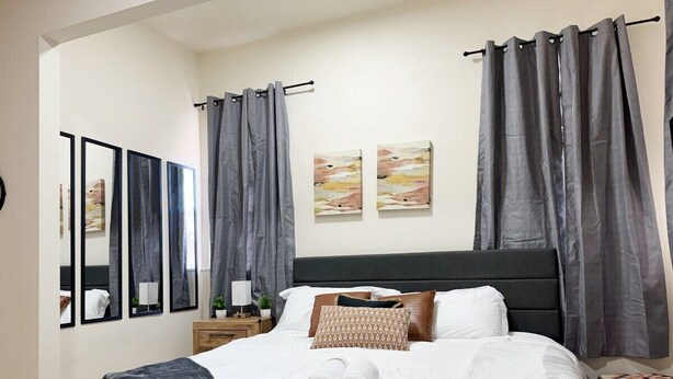 A neatly made bed with white linens and a grey blanket, a wooden nightstand with a lamp, and two framed artworks on the wall. - The Cardinal 808 B - KING 1BR-1BA Apt
 (Evansville)