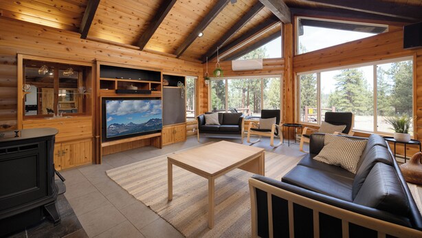 Ein Holzhaus-Interieur mit Holzbalkendecke, Kamin, einer Couch, einem Fernseher und großen Fenstern.