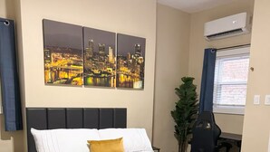 Una cama bien hecha con edredón blanco y una sola almohada amarilla, una obra de arte con un paisaje urbano encima, y un escritorio con una silla a la derecha.