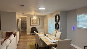 A dining area with a wooden table, chairs, and a rug. There is a ceiling fan and a framed artwork on the wall.