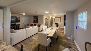A modern living room with a large TV, a dining area with a long table, and a cozy seating area with a sofa and chairs.