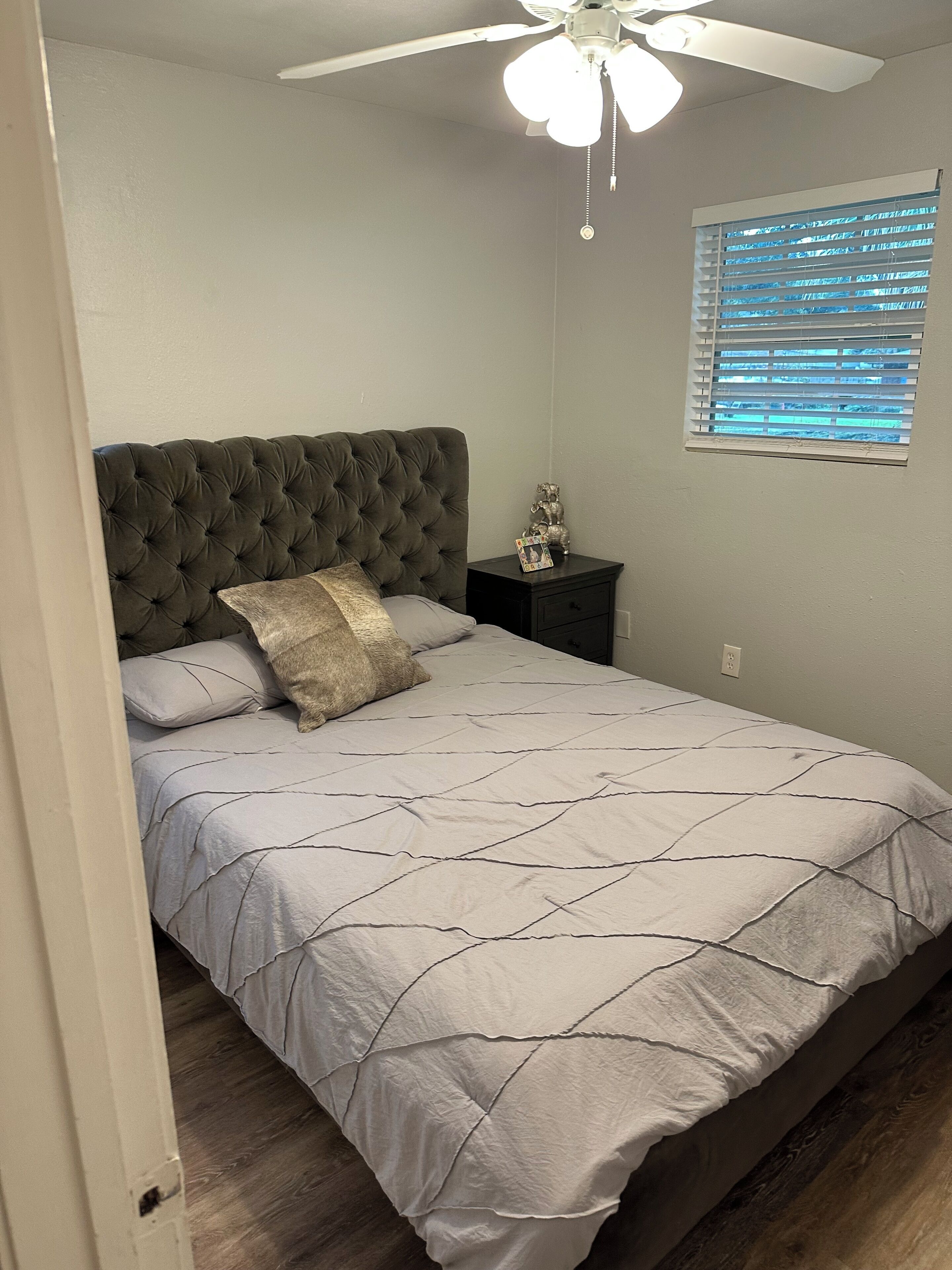 A bedroom with a tufted headboard, a bed with a quilted bedspread, a nightstand with a lamp, and a window with blinds.