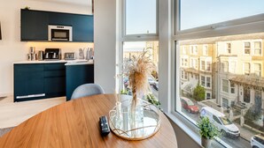 A modern kitchen with dark cabinets and a white countertop, a dining table with a remote control, and a large window with a city view.