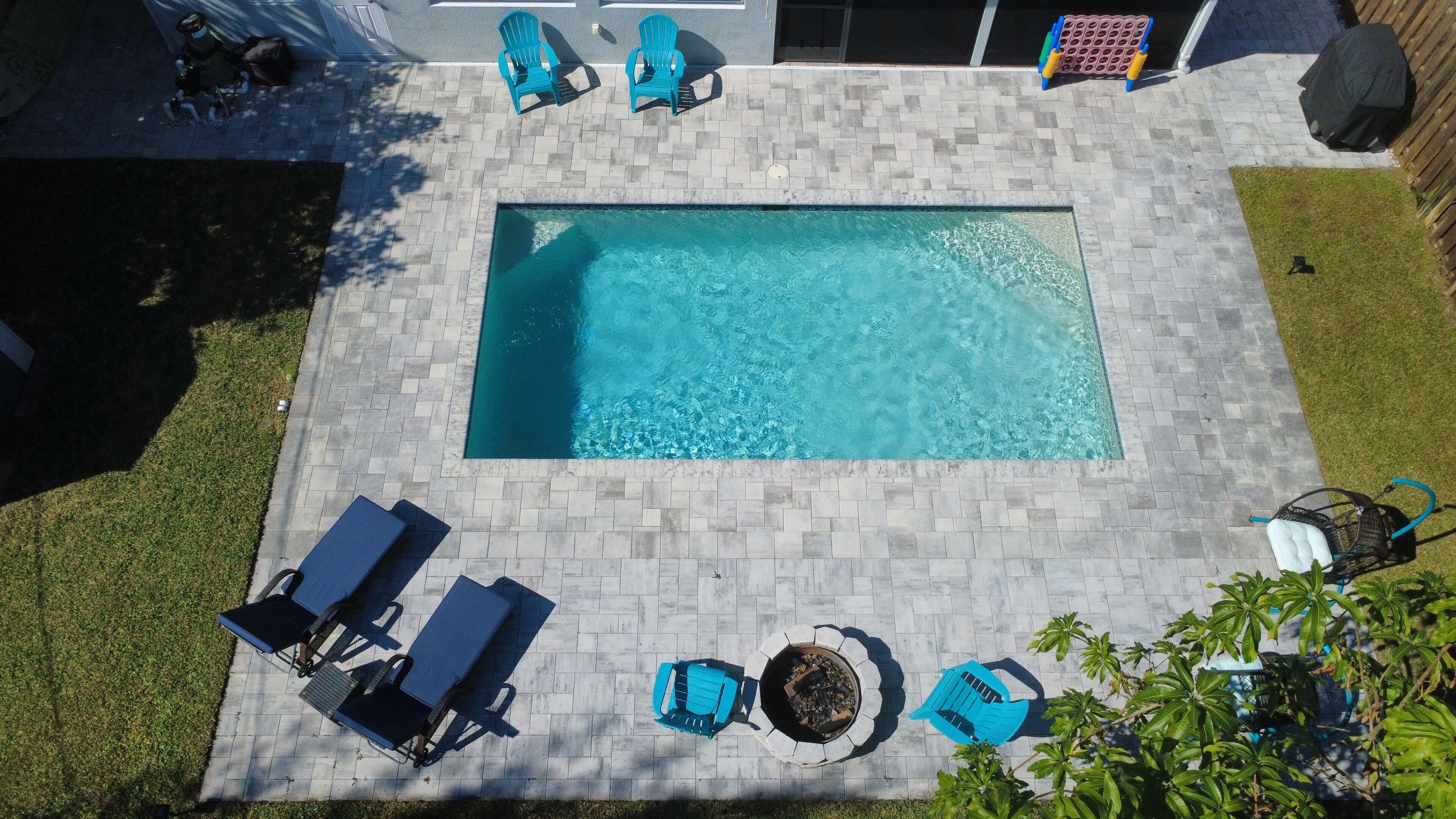 A rectangular swimming pool with a tiled deck, surrounded by blue lounge chairs and a fire pit.
