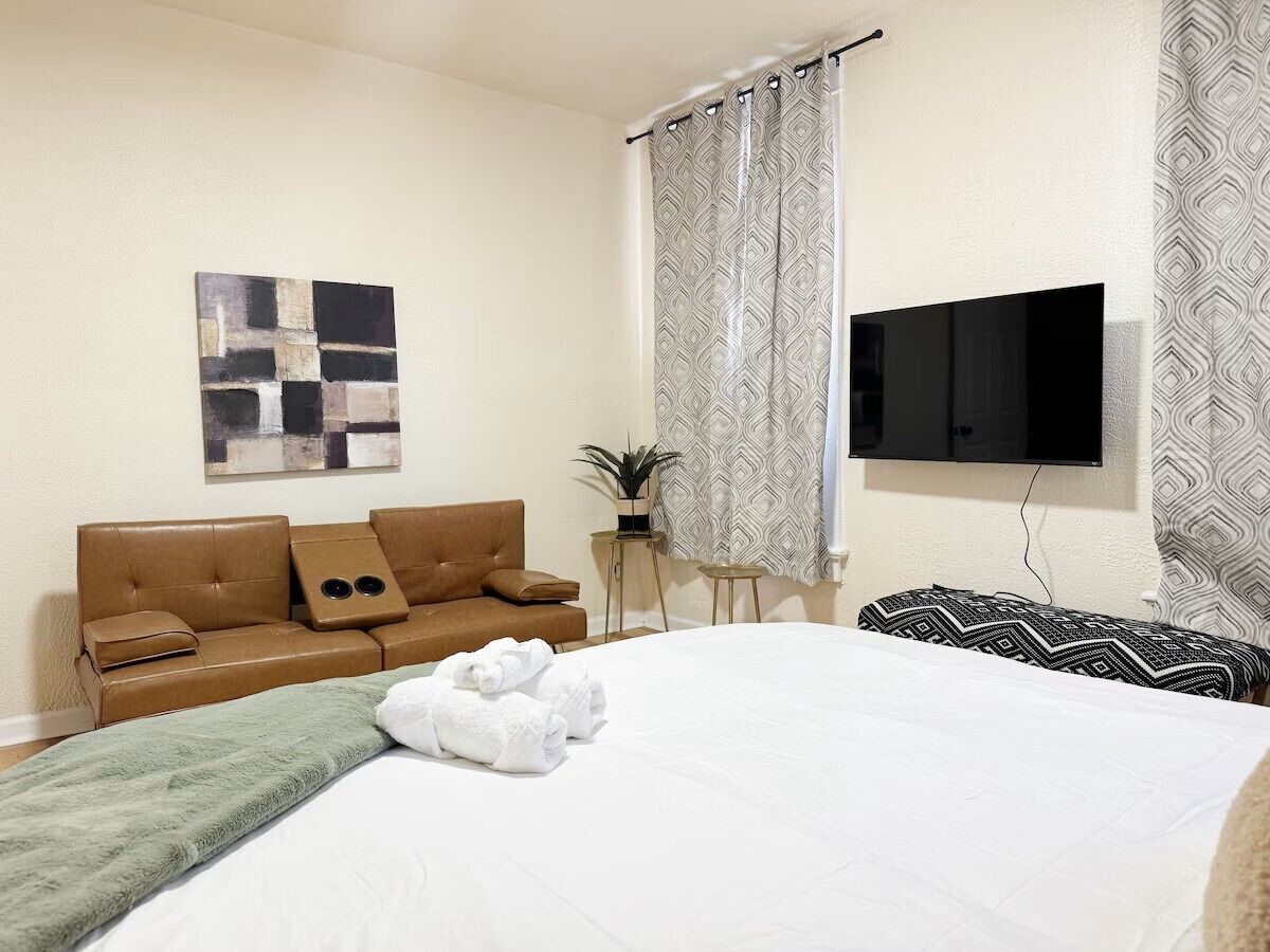 Une chambre d’hôtel avec un lit, un canapé en cuir marron, un téléviseur à écran plat, des rideaux à motifs et une œuvre d’art murale.