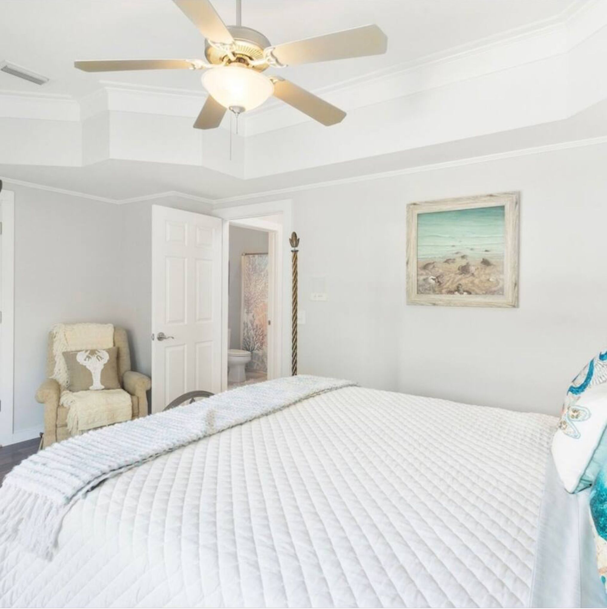 A bedroom with a large bed, a ceiling fan, a door leading to another room, and a framed picture on the wall.
