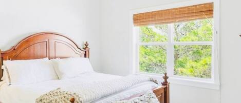 Una camera da letto con un letto di legno, biancheria bianca, una finestra vista sul verde e una ventola a soffitto.