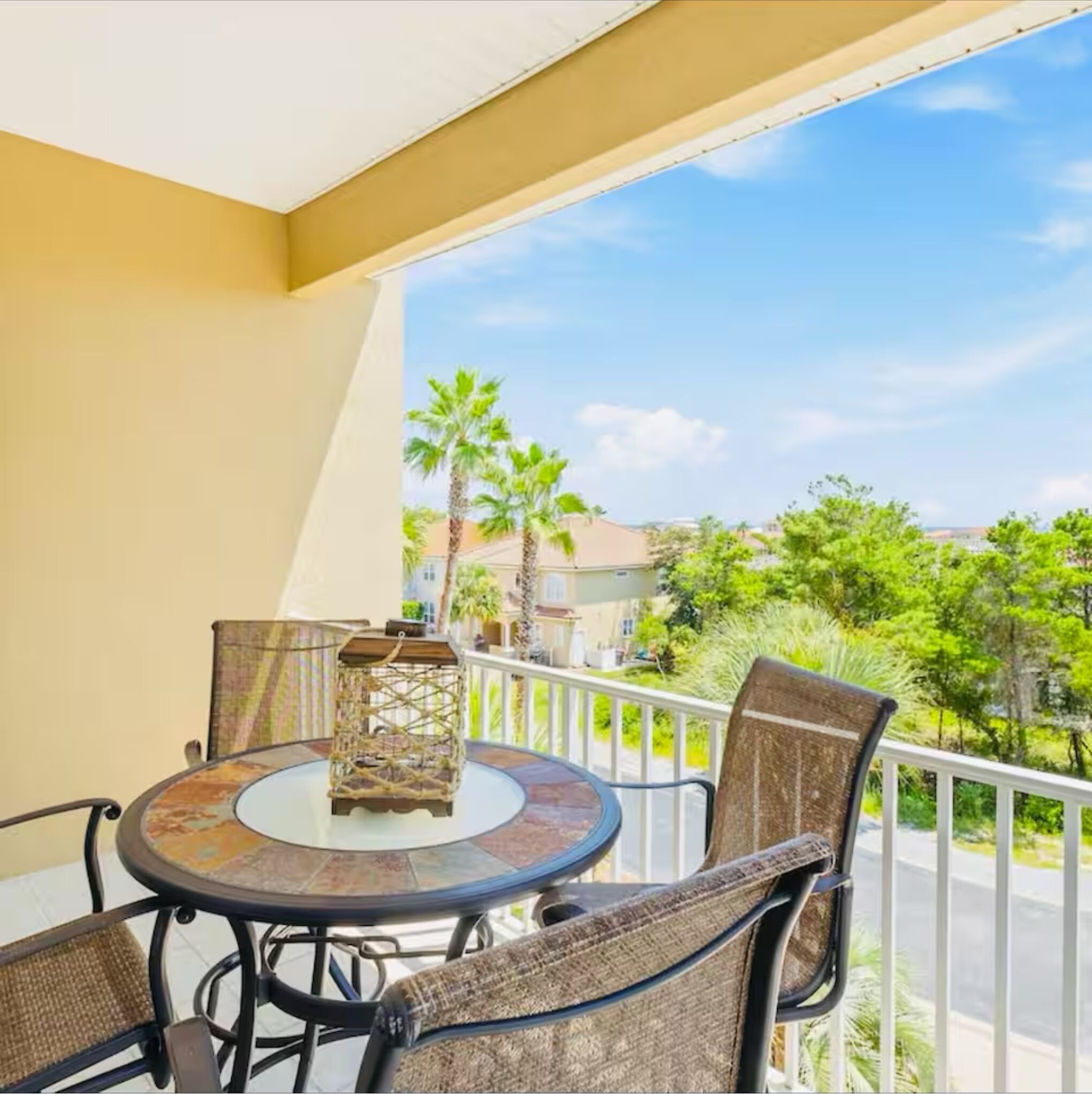 A balcony with a round table and chairs, a decorative lantern, and a view of palm trees and houses.