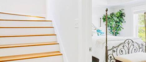 Una escalera con peldaños de madera lleva a un dormitorio con una cama, un ventilador de techo y una ventana con vistas.