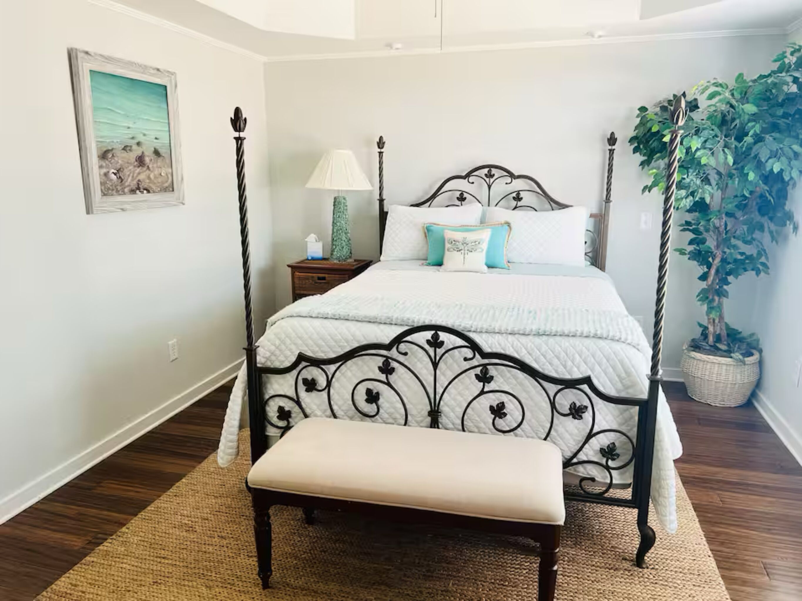 A bedroom with a four-poster bed, a bedside table with a lamp, a bench, and a potted plant.