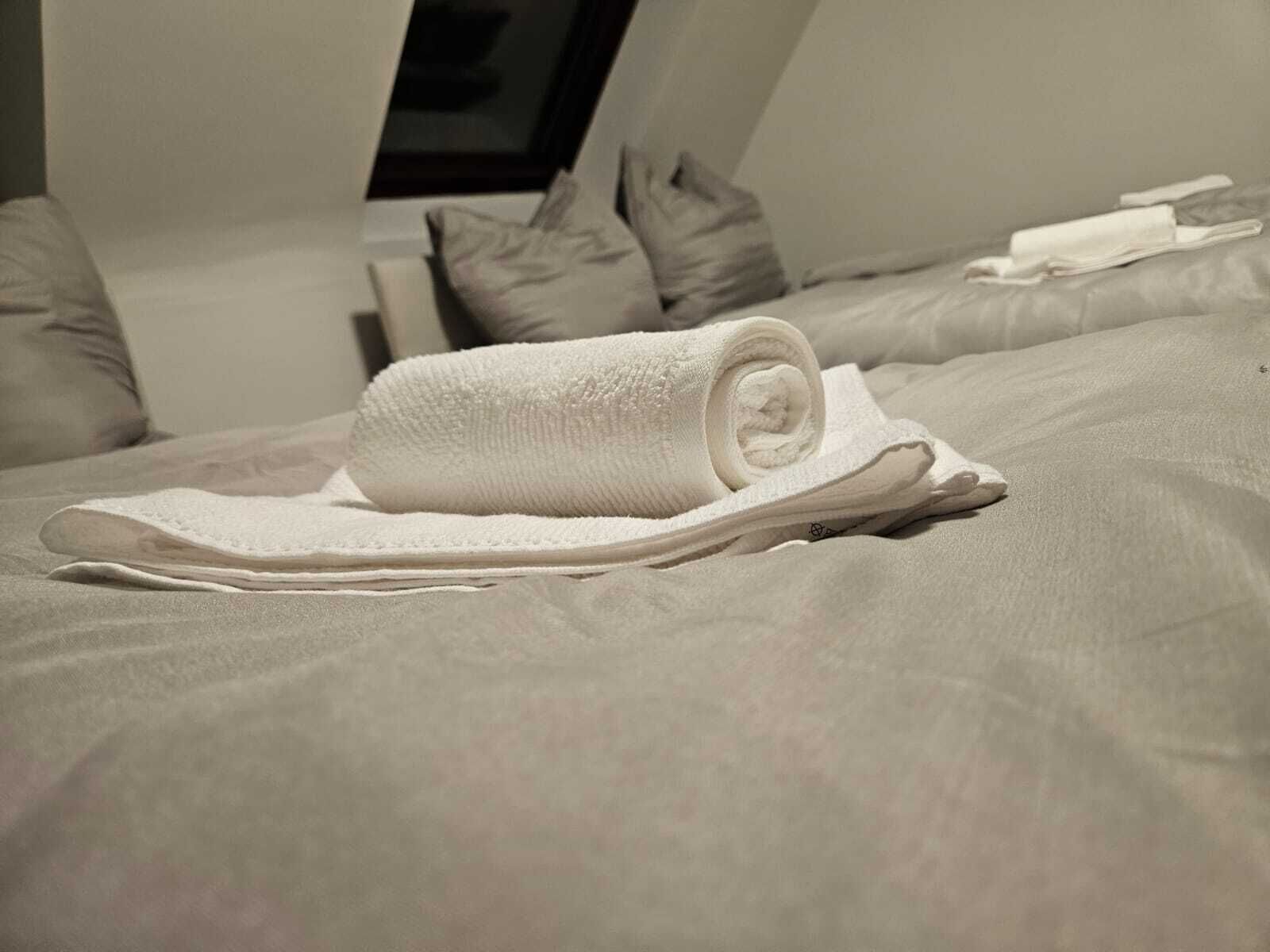 Un letto rifatto con cura, un asciugamano bianco arrotolato e ciabatte, un cuscino e un indumento bianco piegato sul letto.