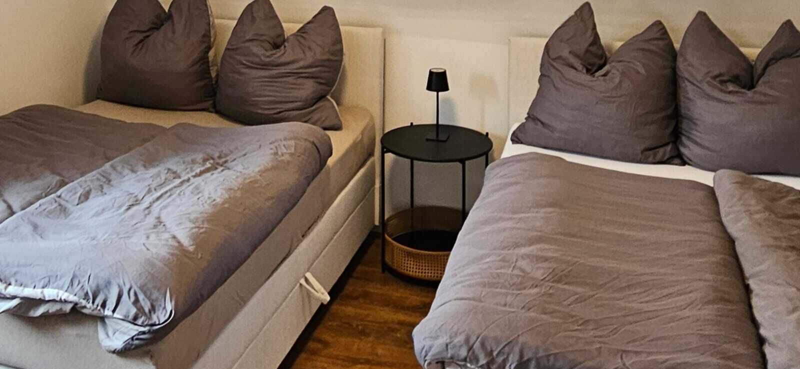 A bedroom with two beds, each with a grey duvet and multiple pillows. There is a small round table with a lamp between the beds.