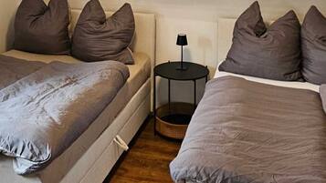A bedroom with two beds, each with a grey duvet and multiple pillows. There is a small round table with a lamp between the beds.