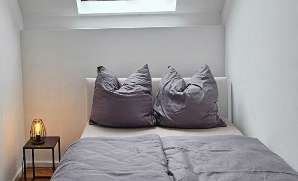 A bedroom with a bed, two pillows, a bedside table with a lamp, and a window above the bed.