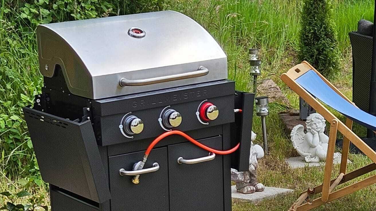 A charcoal grill with a digital display and control knobs, set in a garden with a statue and a sunshade.