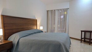 Una cama bien tendida con una colcha estampada, un cabecero de madera, una mesita de noche con una lámpara y una ventana con cortinas ligeras.
