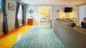Ein modernes Wohnzimmer mit grauem Sofa, einem blauen Teppich mit Muster, Holzboden, einem Fernseher und einem kleinen Beistelltisch mit Lampen.