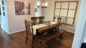 Un coin repas avec une table en bois, des chaises et un banc. Un lustre est suspendu au-dessus de la table et une fenêtre à carreaux se trouve sur le mur.