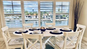A dining area with a round table set for six, white chairs with striped cushions, and a view of a water body through large windows.
