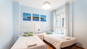 Un dormitorio con dos camas individuales, una mesita de noche con lámpara, una ventana con cortinas y tres cuadros colgados en la pared.