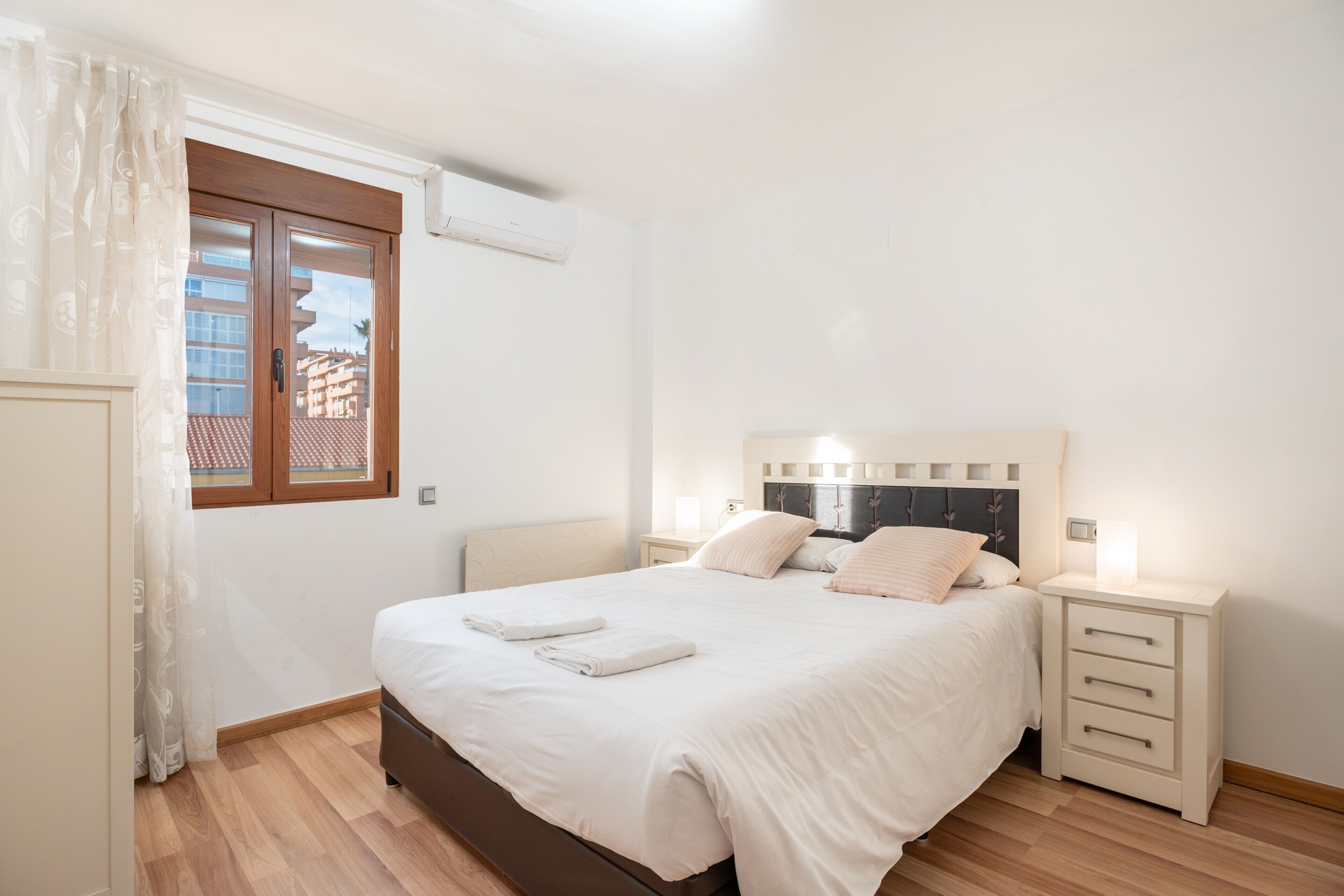 Een slaapkamer met een bed, nachtkastjes, een raam met gordijnen en een airconditioning.