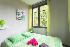 Ein Schlafzimmer mit einem Bett, Kissen, einer grünen Lampe, einem Nachttisch und einem Fenster mit Vorhängen.