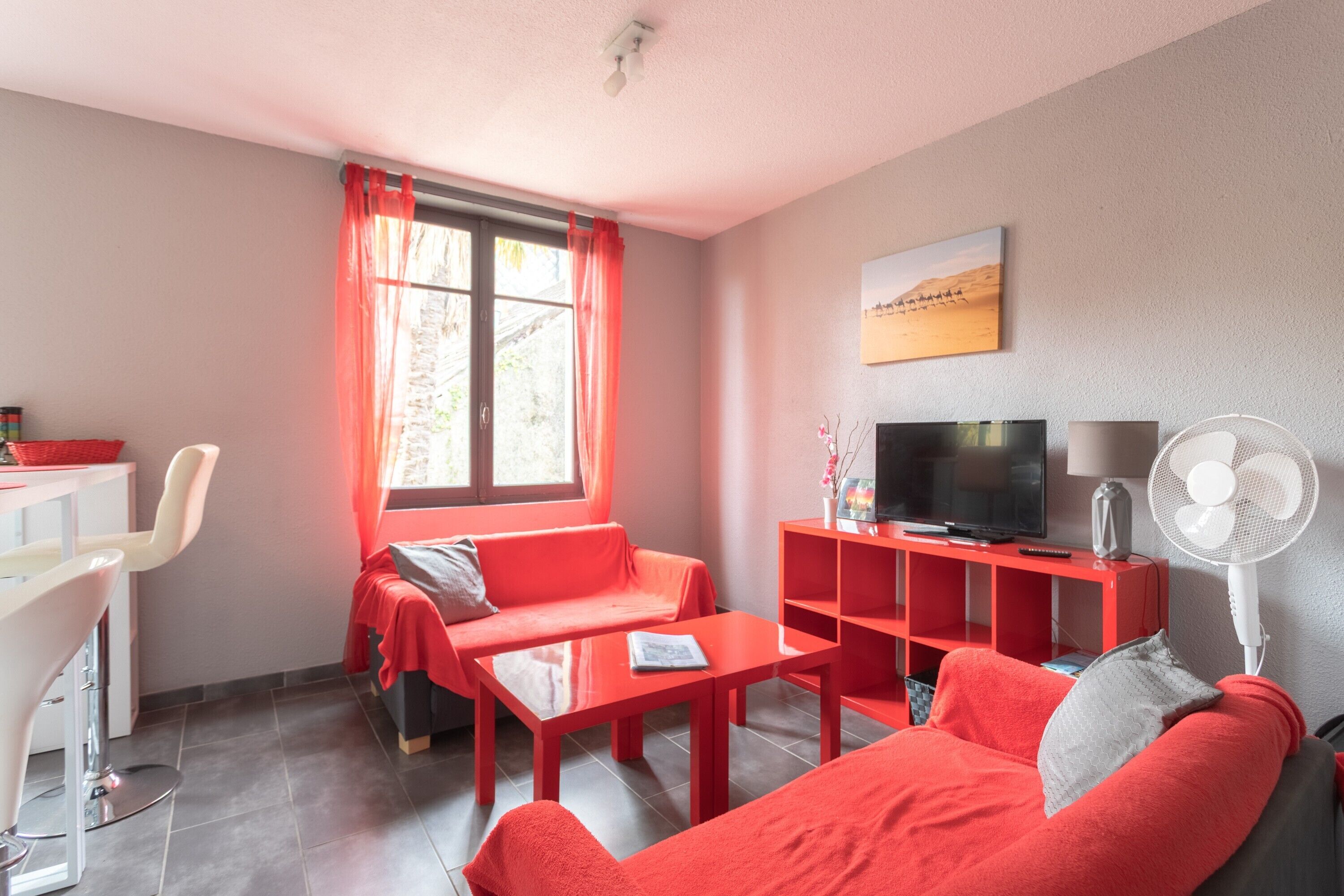 Een moderne woonkamer met een rode bank, een televisie, een witte ventilator en een rode bureau met een stoel.