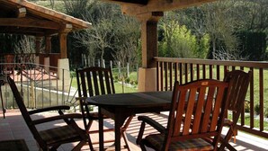 An outdoor patio with a wooden table and chairs, a pergola overhead, and a view of a garden and trees.
