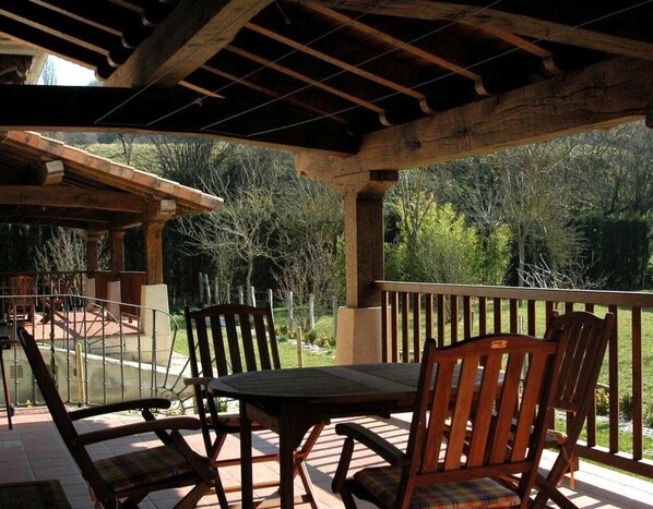 Eine Terrasse im Freien mit einem Holztisch und -stühlen, einer Pergola darüber und Blick auf einen Garten und Bäume.